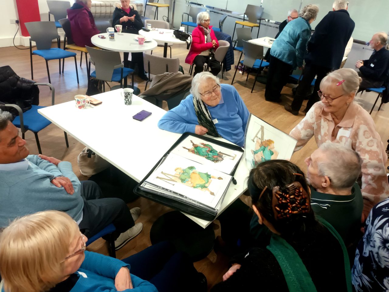 A group of elderly individuals engaging in a social activity in a brightly lit room, with some seated around tables and others standing, while one person showcases artwork in a portfolio for others to view.